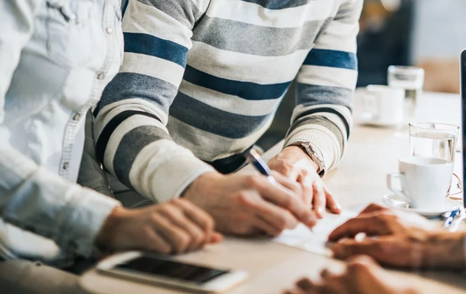 Pessoa assinando um contrato enquanto outra observa atentamente, em um ambiente informal com café sobre a mesa.