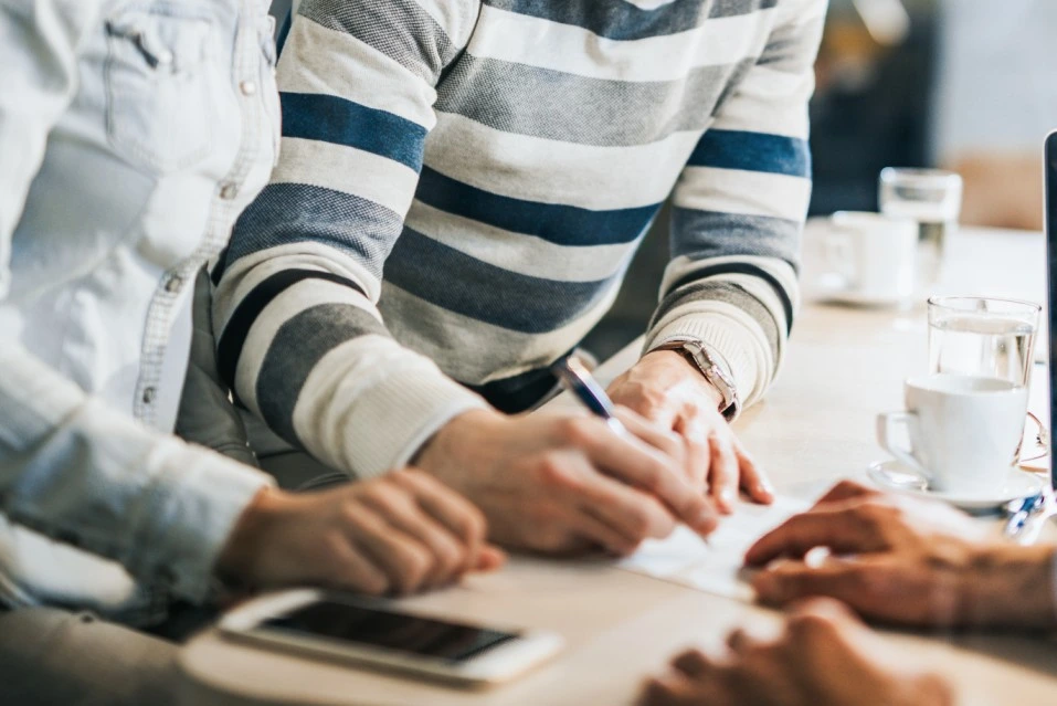 Pessoa assinando um contrato enquanto outra observa atentamente, em um ambiente informal com café sobre a mesa.