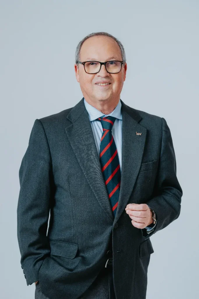 John Redin, consultor imobiliário em Cascais, com traje formal e pin da KW Select, sorridente sobre fundo neutro.