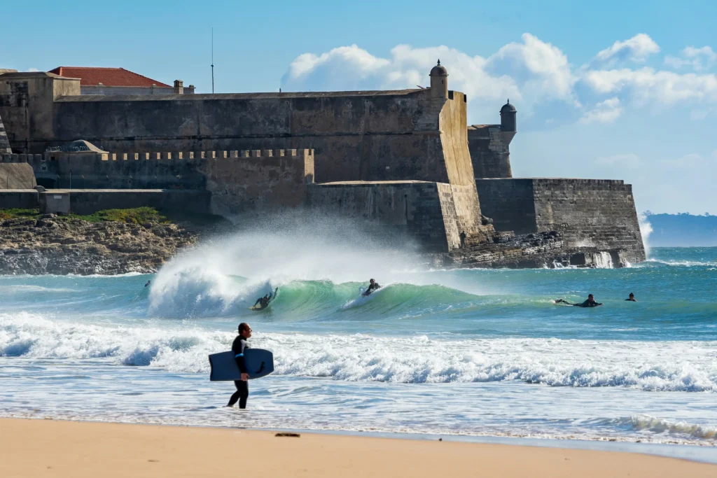 Surfistas a entrar na Praia de Carcavelos com o Forte de São Julião ao fundo, ilustrando o estilo de vida da freguesia.